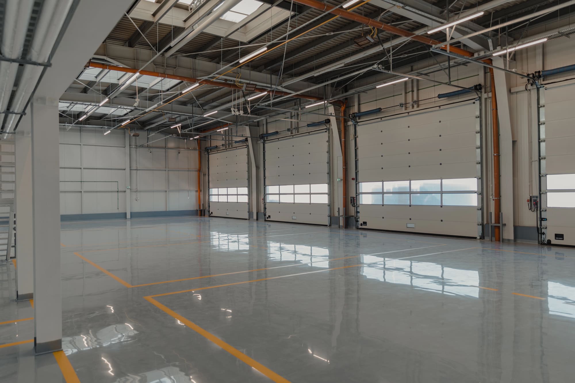 A wide-angle interior view of a professional commercial garage or industrial facility featuring a high-gloss, light-gray epoxy floor coating. The seamless, reflective surface is marked with crisp yellow safety lines and perfectly reflects the overhead industrial lighting and white bay doors. This showroom-quality finish demonstrates the durable and easy-to-clean results provided by Ponce’s Quality Painting Inc. for clients in the Visalia area.