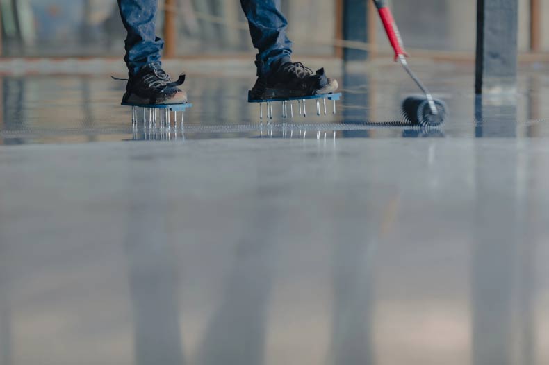 A detailed close-up of a professional installer wearing spiked shoes to walk across a fresh, wet epoxy floor coating without leaving footprints. The specialist is using a spiked roller to smooth the surface and release air bubbles, ensuring a perfectly level, high-gloss finish. This meticulous application process by Ponce’s Quality Painting Inc. demonstrates the precision required for high-quality residential and commercial flooring in the Visalia area.